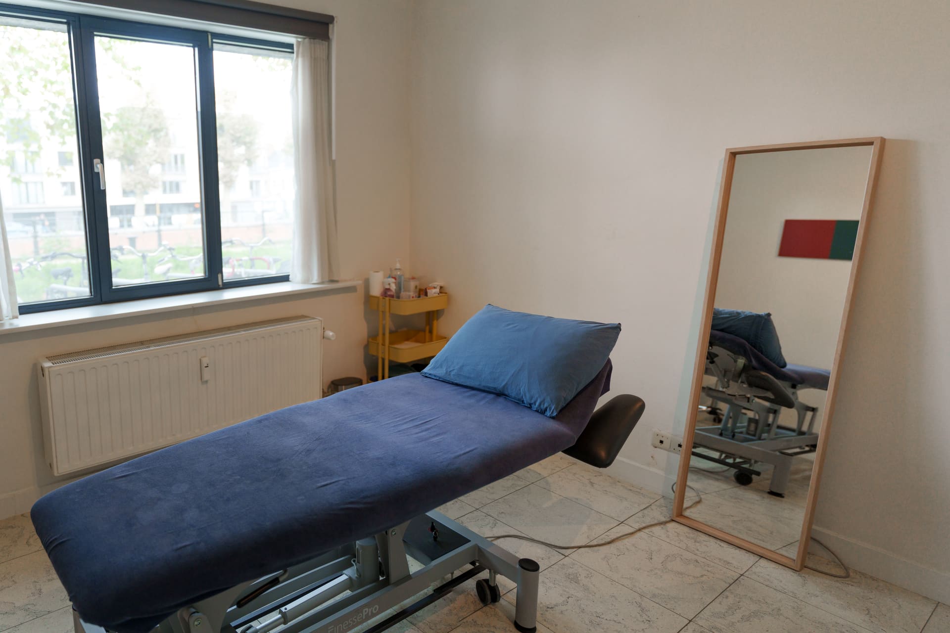 Behandelkamer met blauwe behandeltafel, raam op de straat en een grote spiegel